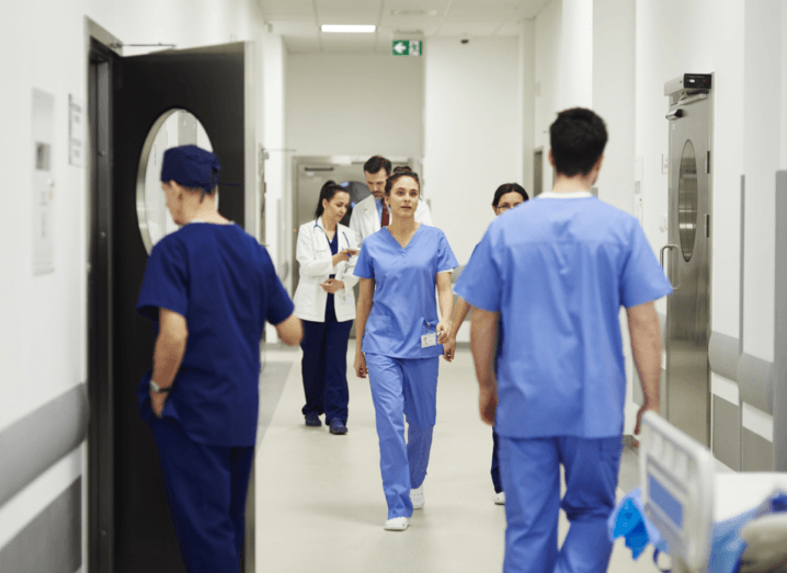 A group of doctors and nurses walking through a hospital corridor. Some are wearing light blue scrubs, others are wearing dark blue scrubs.