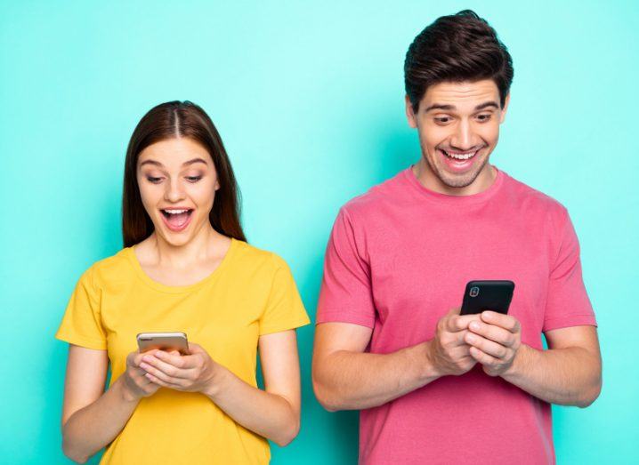 A man and a woman standing side by side wearing colourful T-shirts look down at their smartphones and grin idiotically.