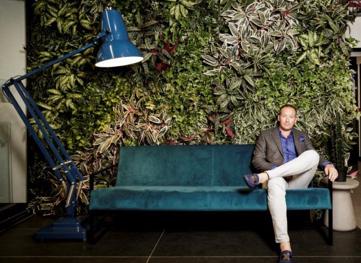 A man in a casual suit sits on a green couch against a floral wall.
