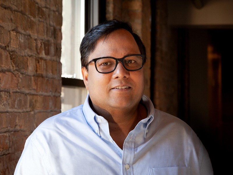 View of man with dark hair and glasses in light-coloured shirt smiling while sitting in front of an exposed-brick wall.