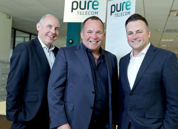 View of three men in business attire standing in front of sign with Pure Telecom logo.