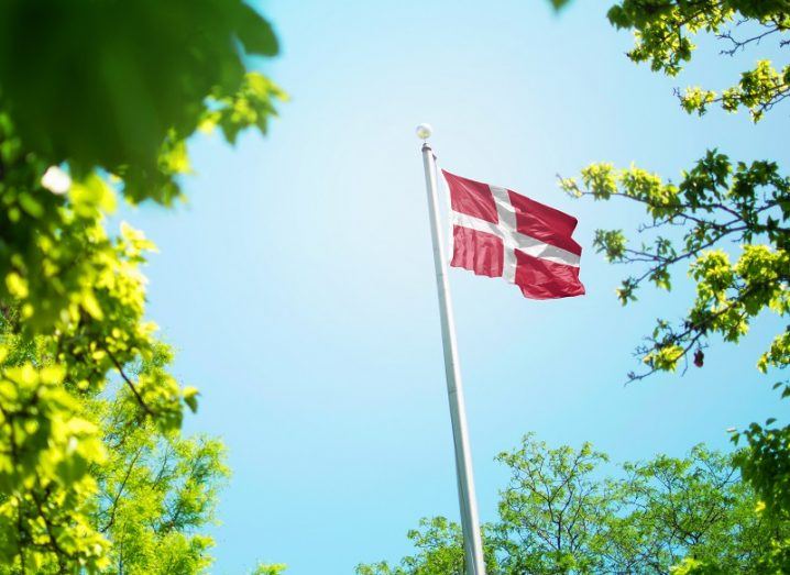 Denmark flag on a pole seen through greenery against a blue sky background.