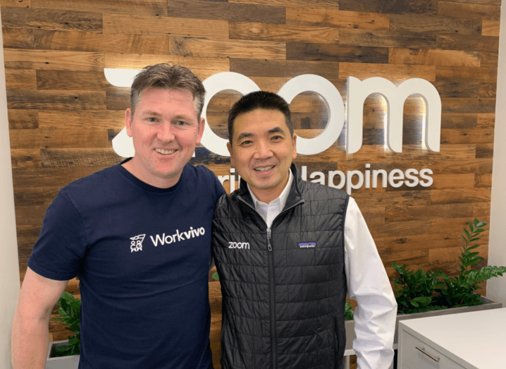 A man with brown hair wearing a navy T-shirt stands beside a man in a white shirt and black Patagonia gilet. The man in the gilet has black hair. They are both standing in front of a large wooden wall that reads "Zoom".