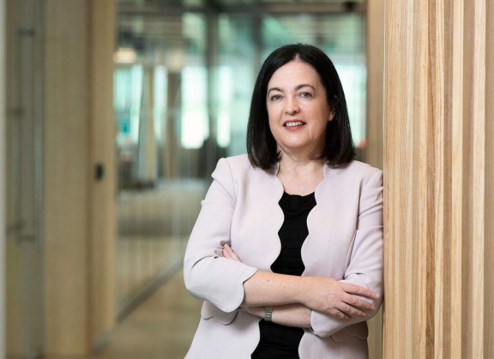 Jacky Fox wears a light scallop-edged blazer, standing with her arms crossed, leaning against a wooden wall in a hallway at Accenture’s Dublin office.