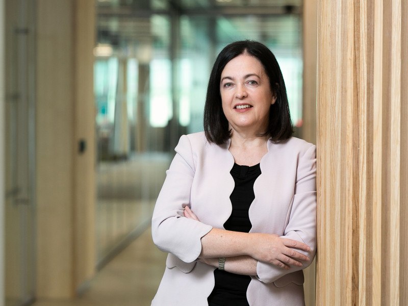 Jacky Fox wears a light scallop-edged blazer, standing with her arms crossed, leaning against a wooden wall in a hallway at Accenture’s Dublin office.