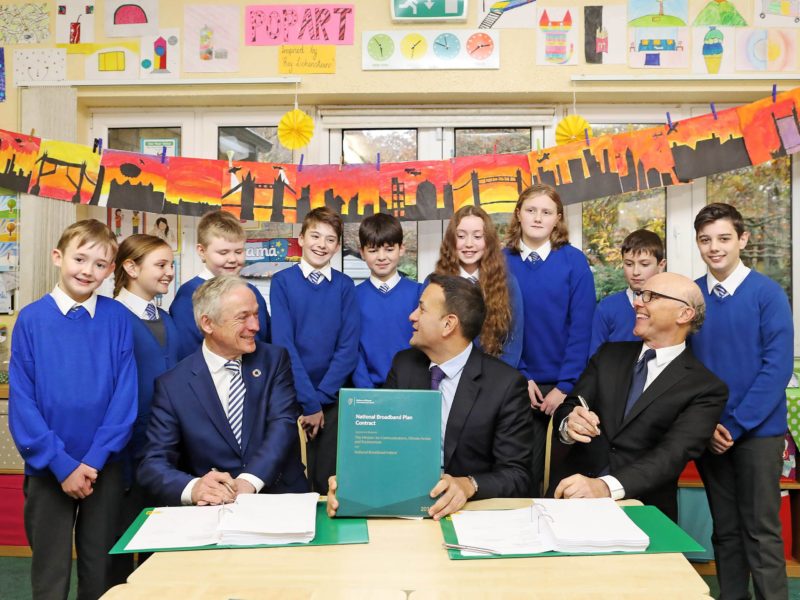 Richard Bruton, Leo Varadkar and David McCourt sitting at a table in a school with a group of children in uniforms behind them. On the table is a copy of the National Broadband Plan.