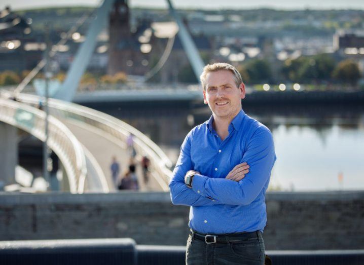 A man in a blue shirt stands with his arms folded in front of a wall, overlooking a bridge.