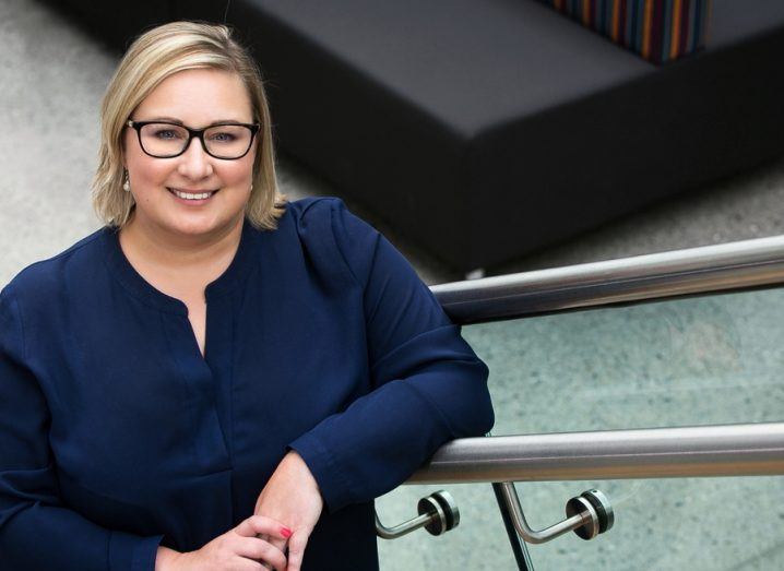 A woman with blonde hair and a dark blue shirt is standing on a staircase and smiling into the camera in a corporate setting at Siemens.