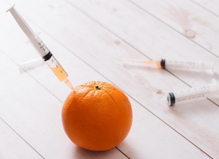 A syringe stuck in an orange on a wooden panelled surface.