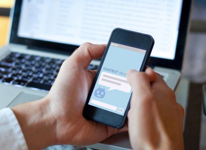 Hand holding a phone an interacting with a chatbot with a laptop in the background.