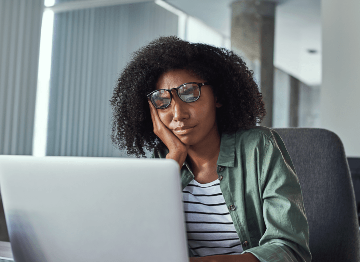 A woman in a green shirt with a white and navy striped T-shirt underneath, sits at a laptop looking frustrated. She is pushing her glasses up with one hand while typing with the other hand. She has black, curly hair.