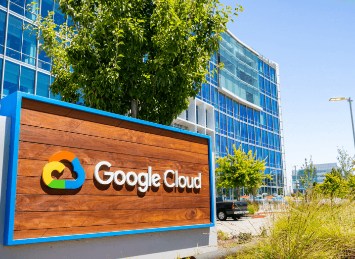 The Google Cloud logo on the outside of a building in California. It is sunny and the building has blue glass. There is a tree above the Google Cloud logo. The logo is yellow, red, blue and green and in the shape of a cloud.