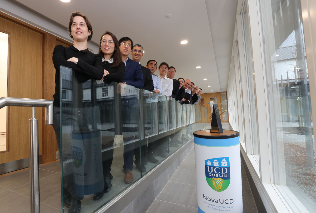 A row of people standing against a glass banister in UCD, posing for a photograph. Beside them is a small podium with the UCD logo on it.