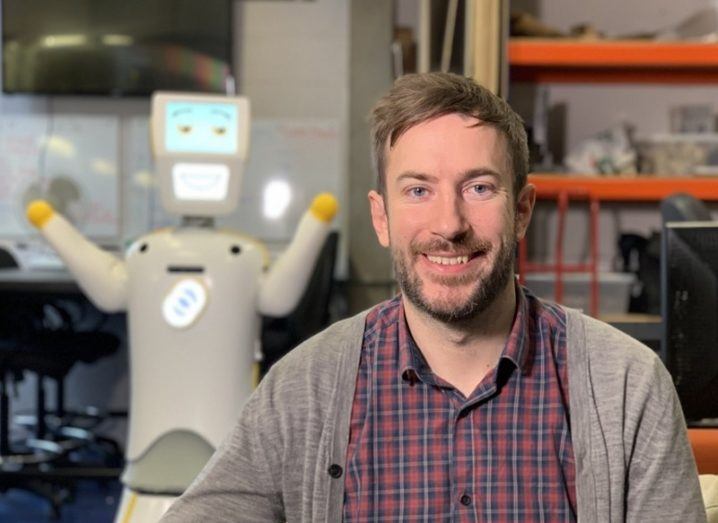 Conor McGinn in a grey cardigan and wine-coloured patterned shirt with the Stevie robot in the background.