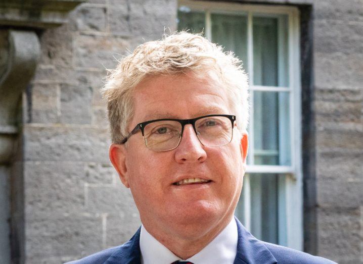 View of blonde man with thick-rimmed glasses in business attire standing in front of stone building.