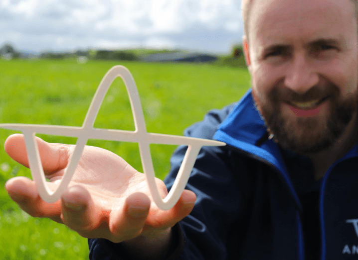 A bearded man with brown hair, wearing a navy fleece, crouches in a field and holds a white 'A' shaped piece of plastic, representing the logo of Anuland.