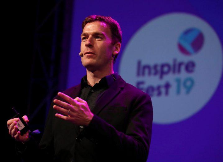 A man in a black suit stands on a stage with a purple screen in the background that reads 'Inspirefest 19'.