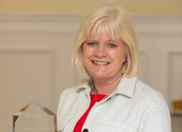 A woman in a white jacket and red top stands in a room holding a glass award.