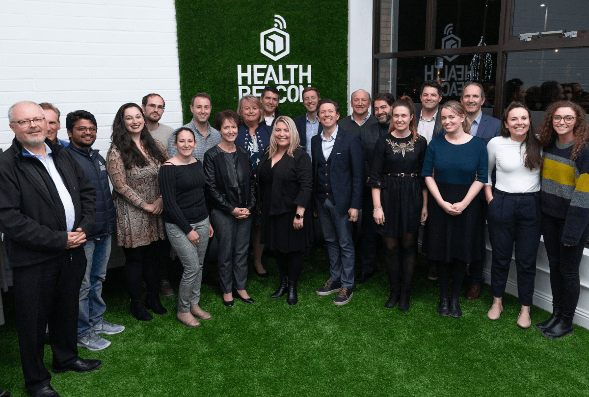 A large group of people standing on a green astroturf rug in front of a white wall that says 'Health Beacon'.