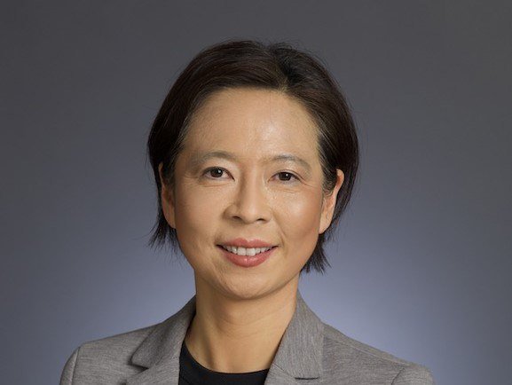 Headshot of woman with sallow skin and cropped black hair in a pixie cut smiling at the camera, wearing a grey blazer.