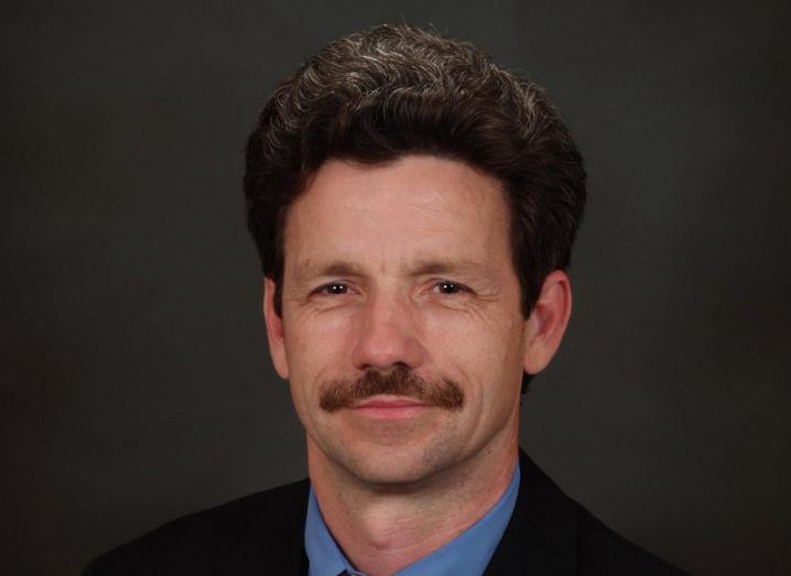 Man with dark hair and moustache looking at camera and smiling against dark background.