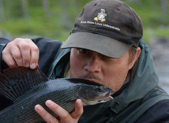 Jens Carlsson in a black baseball cap and rain jacket holding a fish in front of his face.