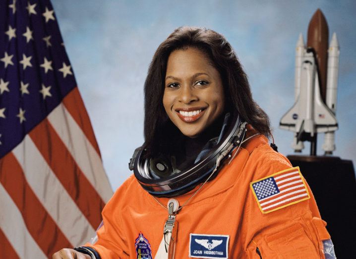A smiling NASA astronaut in an orange flight suit pictured next to an American flag with a picture of a rocket in the background.