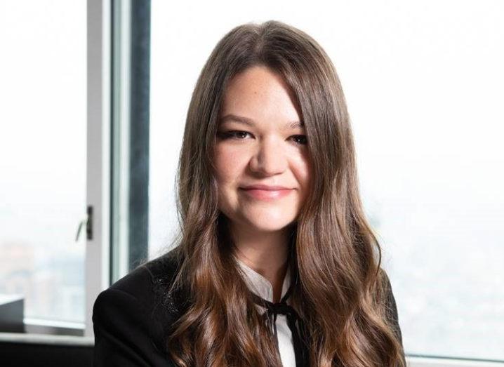 A smiling long-haired woman in a white blouse and black blazer pictured against a window.