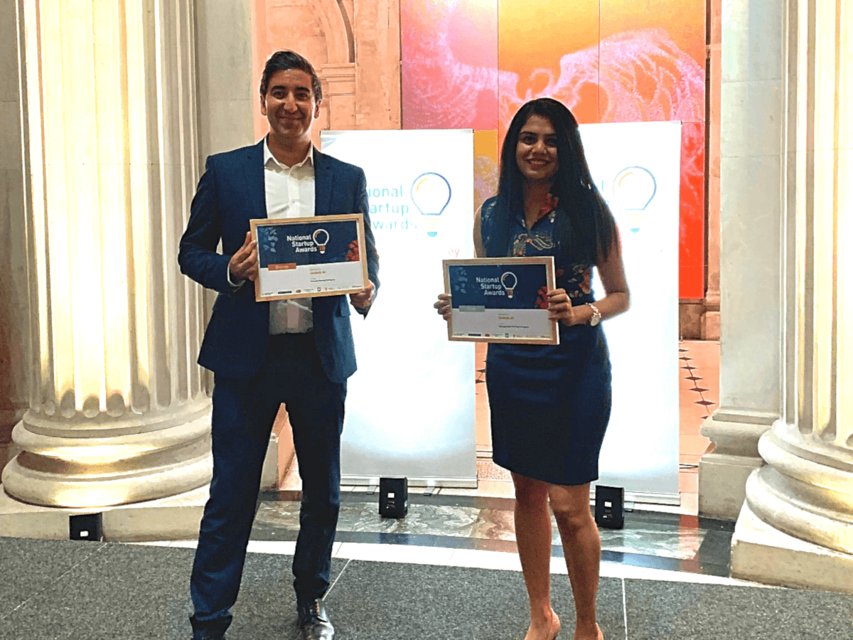 A man in a navy suit and white shirt holds a certificate beside a woman in a navy dress with long, black hair, who is also holding a certificate. They are standing in front of two large, white pillars.