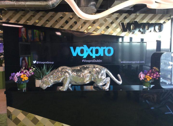 A black reception desk with a large TV screen, which reads 'VoxPro' in blue text. There's a large silver jaguar on the desk of the reception.