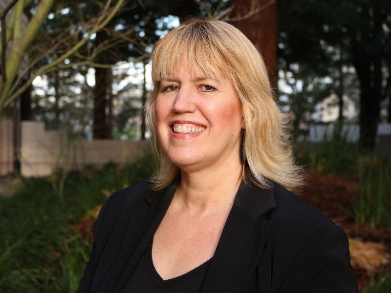 View of blonde woman in dark business attire smiling at camera against forest background.