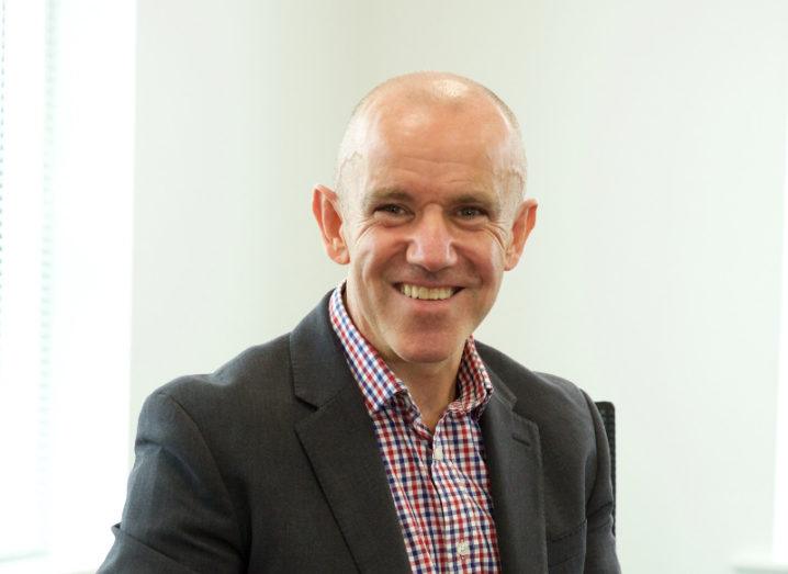 A man in a dark suit and red checkered shirt sits in a bright, white room.