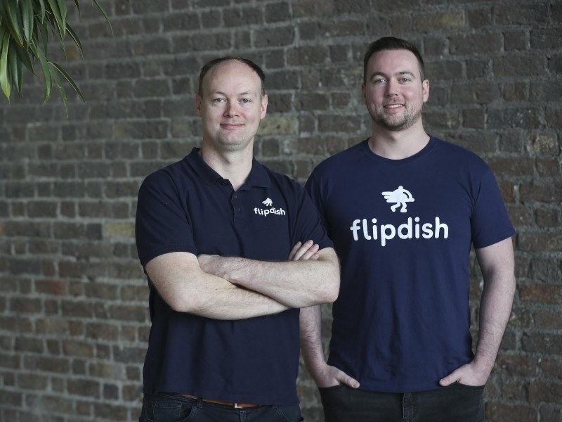 Two men stand in blue 'Flipdish' T-shirts in front of a brick wall.