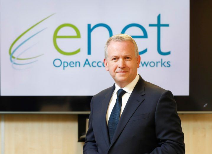 A man in a dark suit stands in front of a screen that says 'enet'.