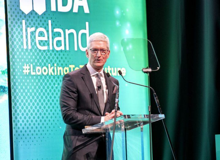 Tim Cook on stage at a podium in the National Concert Hall with a screen behind him bearing the IDA Ireland logo.