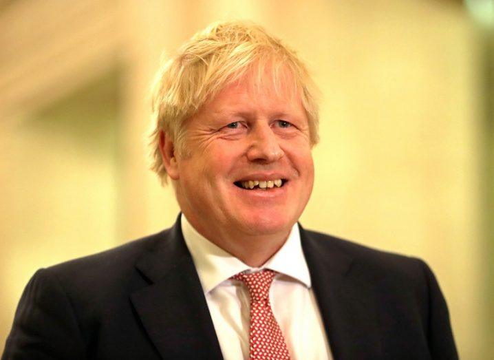 Boris Johnson smiling in a black suit, white shirt and red tie, against a yellow background.