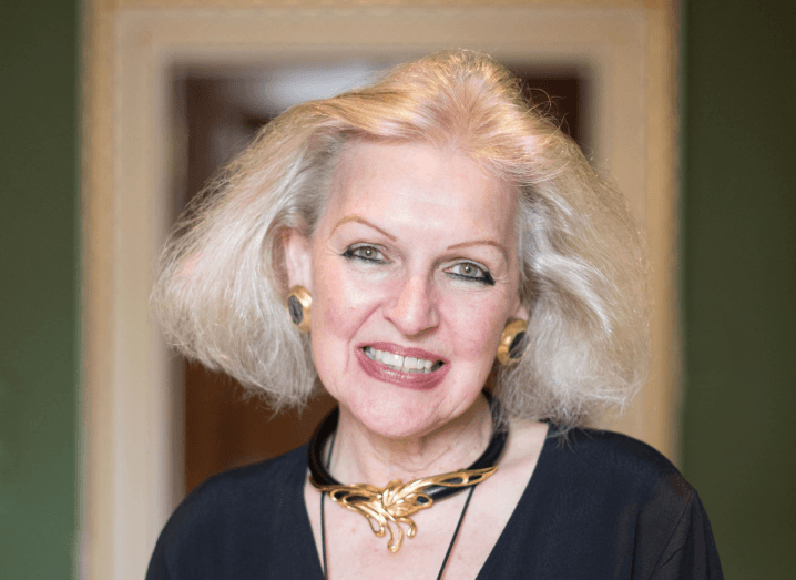 A woman with white hair, large gold earrings and a gold necklace wears a black blouse and stands in front of a green wall.