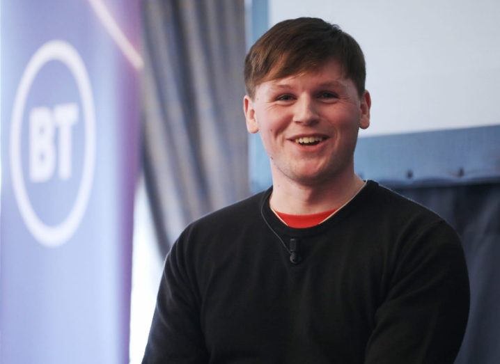 Headshot of a young man smiling broadly and wearing a lapel mic. A BT logo is visible in the background.