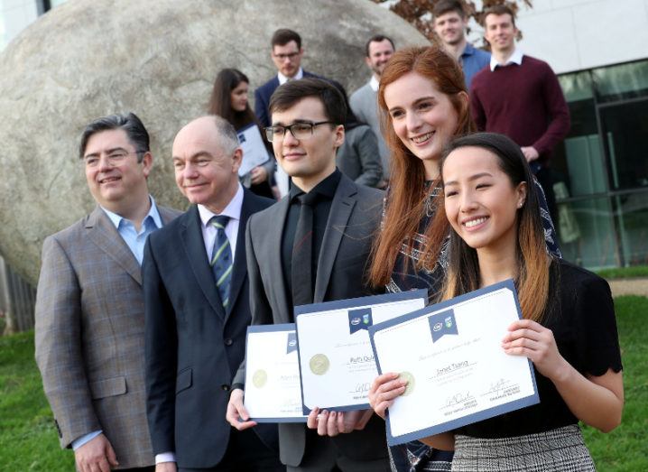 Two men in suits stand among a group of young students holding scholarship certificates on the grounds of a university.