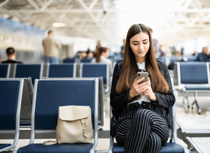 A woman in a black leather jacket and black pinstriped trousers uses a smartphone in an airport terminal. She has long brown hair and a beige bag is placed on the seat beside her.