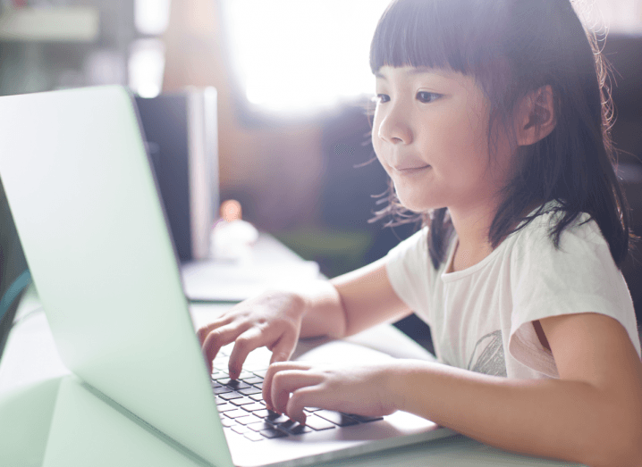 A child typing on a white laptop in a home. She has a white T-shirt and brown hair with a fringe.
