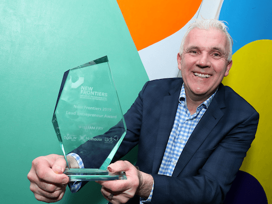 A man in a navy suit with a blue and white shirt holds a glass trophy in front of a mint green wall with orange and white patterns on it.