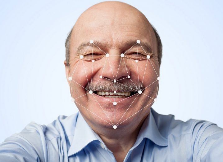 Elderly man with a moustache smiling with facial recognition points rendered over his face against a white background.