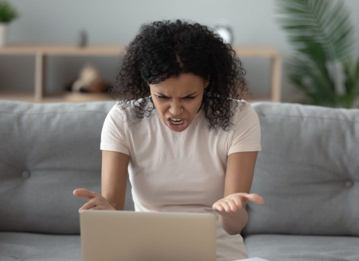 A woman seated on a couch while she works on a laptop contorts her face and gestures at the computer in frustration.