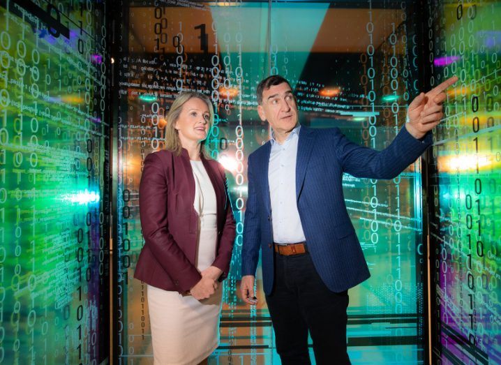 A man and a woman stand in a glass-panelled cubicle with binary code and other text covering the walls. The man points to something out of picture and the woman looks on.