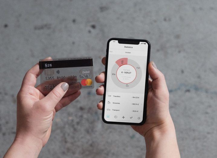 Hands holding an N26 debit card and a phone with the N26 app open, with a concrete floor in the background.