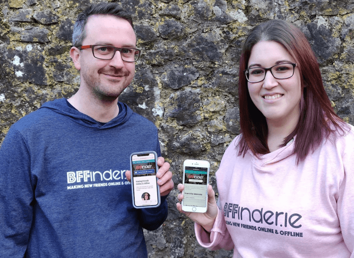 A man in a navy jumper stands beside a woman in a pink jumper. Both of the jumpers are branded with the BFFinder logo. The man has short grey hair and glasses and the woman has red hair.