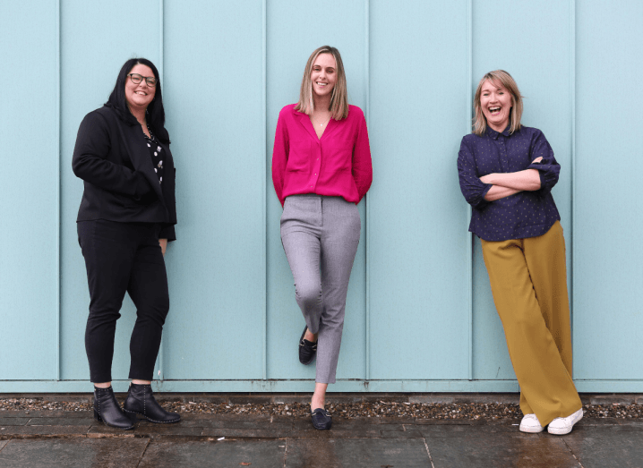 Three women standing in front of a blue wall. The woman on the left is wearing all black and black boots. The woman in the middle is wearing a bright pink cardigan and grey trousers. The woman on the right is wearing a navy blouse and mustard trousers.