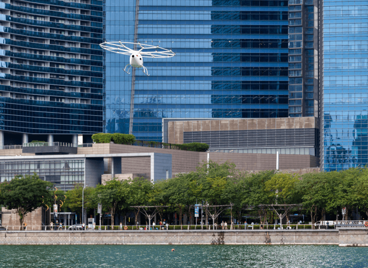 A white aircraft resembling a helicopter flying over a body of water in front of sky scrapers.