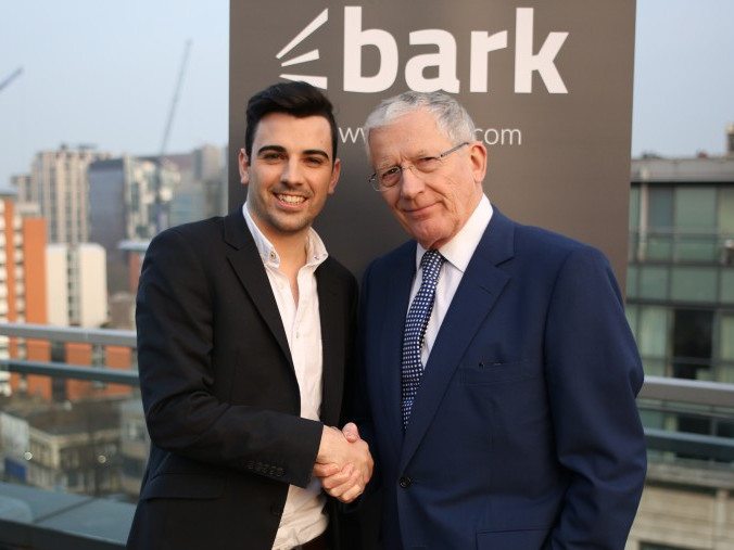 Two men standing beside each other in front of a grey sign that says Bark.